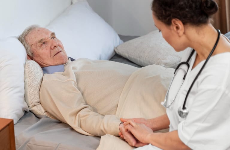 Elderly man laying in bed with a caregiver sitting at the edge of the bed
